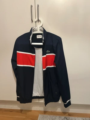 Lacoste dress - Snygg vindjacka från Lacoste i mörkblått med bred röd och vit rand över bröstet och ryggen. Jackan har dragkedja, ståkrage och en röd huva som kan vikas in. Klassisk krokodillogga på bröstet och elastiska muddar vid ärmslut och nederkant. Perfekt för dig som gillar sportig stil.