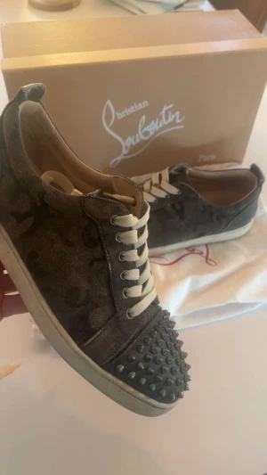 Christian Louboutin camo  - Säljer ett par grymma Christian Louboutin sneakers i mörkgrått camouflagemönster med mocka och skinn. Tåpartiet är täckt av svarta nitar för extra edge. Klassisk röd sula och vita snören. Perfekt för dig som vill sticka ut med lyxig streetstil.