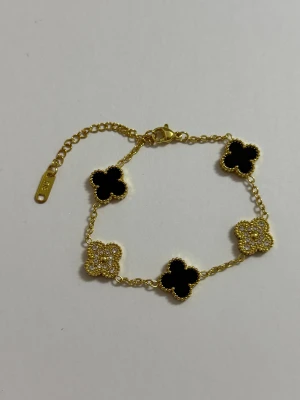 18 k Guldpläterad armband med svarta och glittriga blommor - Snyggt armband i guldfärgad metall med fem fyrklöverformade hängen, tre i svart och två dekorerade med glittrande stenar. Kedjan har en justerbar längd och en liten bricka Perfekt för dig som gillar blommiga detaljer och lite extra bling. 