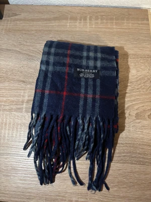 Rutig marinblå halsduk från Burberry - Snygg halsduk från Burberry i marinblått med klassiskt rutmönster i grått och rött. Gjord i mjuk kashmir, med fransar i ändarna och diskret Burberry-logga. Perfekt för att ge din outfit en cool och lyxig touch.