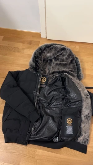 Svart pufferjacka Glorious Gangsta  - Svart pufferjacka från Glorious Gangsta med fluffig grå fuskpäls i luvan, snygga detaljer som guldiga dragkedjor och en stor ficka på ärmen. Jackan har quiltad design, knappar framtill och är fodrad med mönstrat tyg. Perfekt för kalla dagar med street vibe.