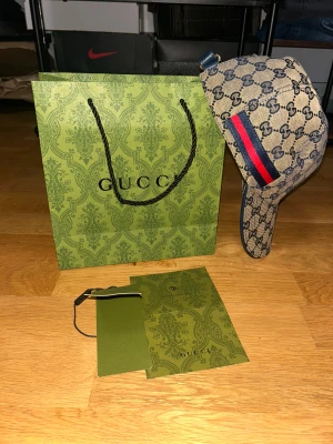 Gucci keps  - Exklusiv Gucci keps i beige med klassiskt GG-monogram i blått, röd-blå rand på sidan och justerbart blått läderband bak. Tillverkad i 100% bomull och läderdetaljer. Säljes med originalpåse och tags. Perfekt för dig som vill sticka ut med lyxig streetstyle.