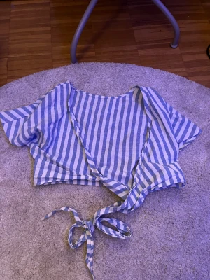 Randig omlottopp från SHEIN, S - Säljer en fräsch omlottopp från SHEIN i storlek S. Toppen är croppad med korta ärmar och har blåvita vertikala ränder. Knytband framtill för en snygg passform. Materialet är lätt och luftigt, perfekt för varma dagar.