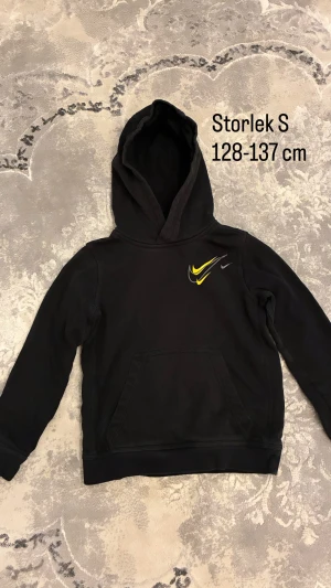 Svart Nike hoodie med gul logga - Svart hoodie från Nike i storlek S med klassisk huva och magficka. På bröstet finns tre Nike-loggor i gul, grå och silver. Perfekt för dig som gillar sportig stil och vill ha något bekvämt och snyggt.