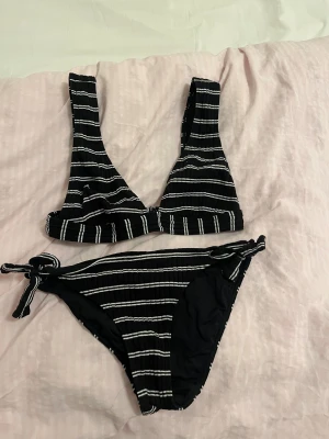 Svart och vit randig bikini med knyt - Snygg bikini med svart bas och vita ränder. Toppen har breda axelband och spänne i ryggen, medan bikinitrosan har knyt på sidorna för en justerbar passform. Materialet är ribbat.