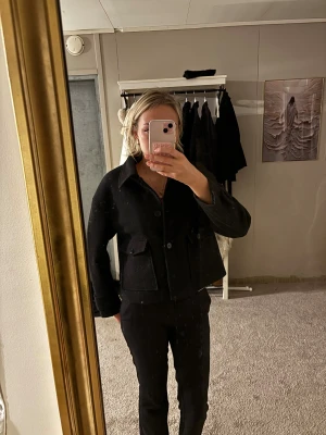 Svart kort kappa - Säljer en svart, kort kappa med klassisk krage och stora fickor framtill. Jackan har knappar hela vägen och är i ett tjockare ullmaterial som håller dig varm. Snygg loose passform och stilren look som funkar till många outfits.