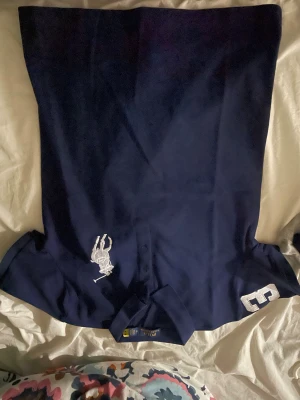 Mörkblå Polo Ralph Lauren skjorta - Snygg mörkblå skjorta från Polo Ralph Lauren med klassisk krage och vit broderad logga på bröstet. På ena ärmen finns en stor vit siffra 3 som ger en sportig vibe. Perfekt för dig som gillar stilrena och ikoniska plagg.