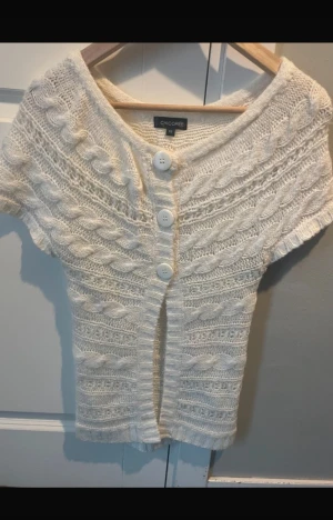 Vit stickad kofta från Chicorée XS - Supersöt vit stickad kofta från Chicorée i storlek XS. Koftan har korta ärmar, grova flätstickade detaljer och tre stora vita knappar framtill. Perfekt för dig som gillar mysiga och trendiga plagg med lite vintagekänsla.