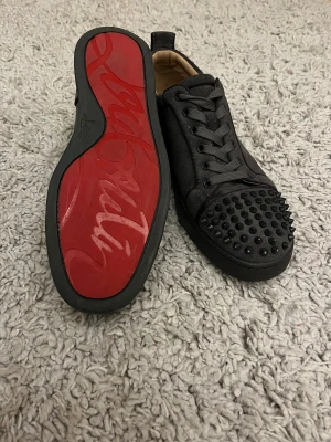 Gråa Louboutins med nitar - Säljer ett par gråa sneakers från Christian Louboutin med ikonisk röd sula och snörning. Skorna har rund tå och är dekorerade med snygga nitar på tån för en edgy look. Ovandelen är i mocka och insidan är ljusbeige. Perfekt för dig som gillar statement pieces. De har små limrester som kan ses på bild 4 men det är inget som syns när man använder skorna! Pris kan diskuteras vid snabb affär!