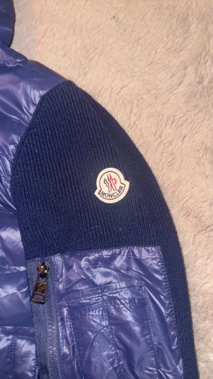 Mörkblå pufferjacka från Moncler - Snygg mörkblå pufferjacka från Moncler med stickade ärmar och sidopaneler. Jackan har huva, dragkedja framtill och Moncler-logga på ärmen. Materialmix med polyester och stickad ull ger en cool look. Fickor med dragkedja och tryckknappar på framsidan. Ena knappen är sönder samt så funkar inte nfc