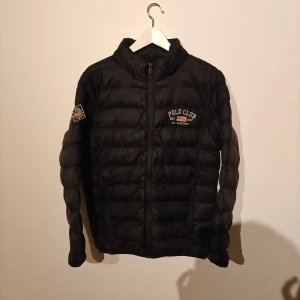 Polo Club vinterjacka - Svart pufferjacka från Polo Club Saint Vincent med broderad logga på bröstet och ärmen. Jackan har huva med tryckt text, dragkedja framtill och två sidofickor. Tillverkad i 100% nylon, perfekt för kalla dagar. Snygg, sportig look med quiltad design.