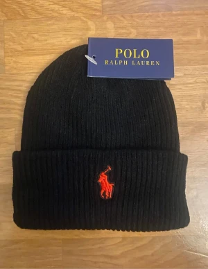 Svart ribbad mössa från Polo Ralph Lauren - Svart ribbstickad mössa från Polo Ralph Lauren med klassisk uppvikt kant och röd broderad logga framtill. Perfekt för kalla dagar och enkel att matcha med din stil. Snygg och tidlös design som passar alla.