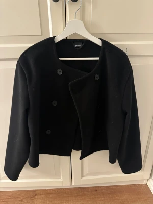 Svart kavajjacka från Gina Tricot - Säljer en svart, stilren kavajjacka från Gina Tricot i storlek S. Jackan har rundad hals, dubbelknäppning med stora knappar och är i en croppad modell.
