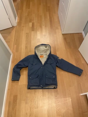 Blå parkajacka från Fjällräven S - Säljer en blå parkajacka från Fjällräven med mysigt teddyfoder och huva. Jackan har flera praktiska fickor framtill, knappar och dragkedja. Perfekt för kyliga dagar och outdoor vibes.