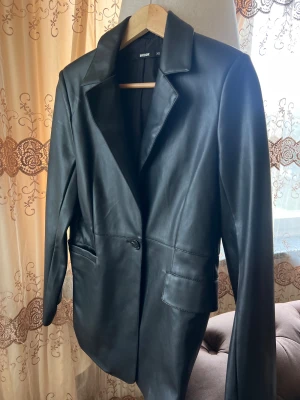 Svart skinnkavaj från Bik Bok XS - Snygg svart kavaj i skinnimitation från Bik Bok, storlek XS. Jackan har klassisk krage, en knapp framtill och två fickor med lock. Stilren och edgy look som passar perfekt till jeans eller kjol. 