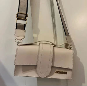 Beige axelväska  - Snygg beige axelväska med stilren design och guldfärgad logga. Väskan har ett brett justerbart axelband i textil och ett handtag upptill. Tillverkad i slätt skinn med dekorativa sömmar och praktiskt lock.