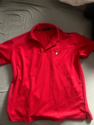 Röd pikétröja från Lyle & Scott - Snygg röd pikétröja från Lyle & Scott med klassisk krage och korta ärmar. Tröjan har det ikoniska gula fågelmärket broderat på bröstet och är gjord i mjuk bomull. Perfekt för dig som gillar stilrena och sportiga plagg.