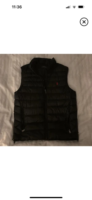Svart dunväst från Polo Ralph Lauren XS - Svart quiltad dunväst från Polo Ralph Lauren i storlek XS. Västen har dragkedja framtill, två sidofickor med dragkedja och röd broderad logga på bröstet. Perfekt att slänga över en hoodie för extra värme. Snygg och stilren design.