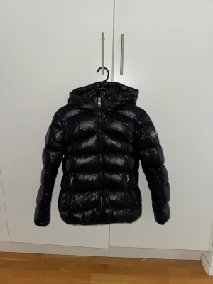 Svart dunjacka från Moncler, 14Y - Snygg svart dunjacka från Moncler med glansig finish och huva. Jackan har två fickor med dragkedja, Moncler-logga på ärmen och quiltad design. Perfekt för kalla dagar, med lätt och fluffig känsla tack vare äkta dunfyllning.