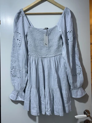 Ljusblå klänning från Bik Bok - Supersöt ljusblå kortklänning från Bik Bok med smockad överdel och volangkjol. Klänningen har långa ärmar med broderade detaljer och lätt puff vid axlarna. Tillverkad i mjukt bomullstyg, perfekt för vår och sommar. Säljes med tag kvar!