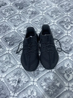 Svarta Adidas Yeezy Boost 350 sneakers - Säljer ett par svarta Adidas Yeezy Boost 350 sneakers med stickad ovandel och platt sula. Skorna har snörning och en diskret röd detalj på hälen. Den stickade strukturen ger en modern vibe och sulan är designad för extra komfort. Perfekt för dig som gillar streetwear och vill ha något stilrent.