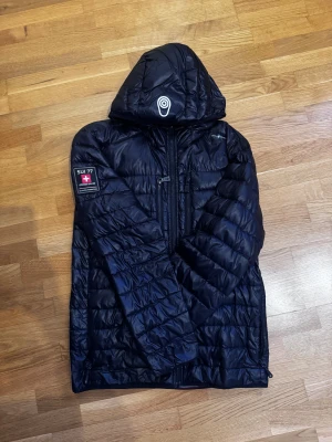 Svart pufferjacka från Sail Racing - Svart pufferjacka från Sail Racing med huva och snyggt SUI 77 Extreme Sailing-märke på ärmen. Jackan har quiltad design, dragkedja framtill och är tillverkad i glansig syntet. Perfekt för dig som gillar sportig stil och vill hålla dig varm under höst och vinter. Det finns ett litet hål som är igen tejpat men inget man ser och ett del på dragkedjan saknas men den går ändå lätt att öppna och stänga. 