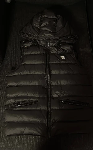 Bvaldi väst  - Svart dunväst från Bvaldi med avtagbar huva och snyggt logomärke på bröstet. Västen har två dragkedjeförsedda fickor framtill och quiltad design med breda horisontella kanaler. Perfekt för lager-på-lager och streetstyle.