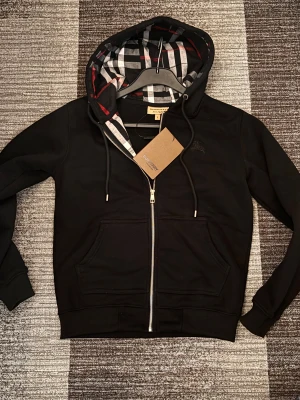 Svart Burberry kofta - Svart hoodie från Burberry med klassiskt rutigt foder i huvan och broderad logga på bröstet. Tröjan har dragkedja, snörning vid huvan och två stora fickor framtill. Tillverkad i mjuk bomull för extra komfort.