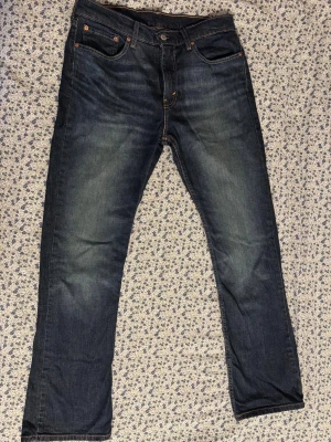 Levi's 527 mörkblå jeans W32 L34: 6 månader gamla - Säljer ett par Levi's 527 jeans i mörkblå tvätt med klassisk femficksdesign och snygga slitningar. Jeansen har raka ben och orangea sömmar, tillverkade i robust denim. Perfekta för dig som gillar en tidlös och stilren look.
