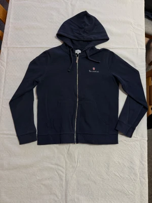 Mörkblå hoodie från Bondelid med guldzip - Snygg mörkblå hoodie från Bondelid i storlek M. Tröjan har huva med snörning, guldigt dragkedjespänne och broderad Bondelid-logga med röd/vit sköld på bröstet. Tillverkad i mjuk bomull och har två fickor framtill. Perfekt för chill och streetstyle.