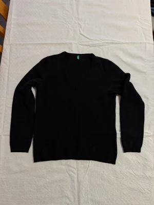 Svart v-ringad tröja från Benetton - Svart v-ringad tröja från United Colors of Benetton i storlek L. Tröjan är stickad i mjuk ull och har långa ärmar samt en diskret broderad logga vid nederkanten. Perfekt för dig som gillar enkel och stilren look.