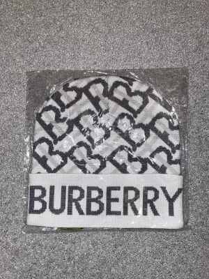 Vit Burberry mössa med logga - Snygg vit mössa från Burberry med grått mönster av B-bokstäver och stor Burberry-logga framtill. Mössan är stickad och har en uppvikt kant. Perfekt för dig som vill ha en lyxig och trendig accessoar till vintern.