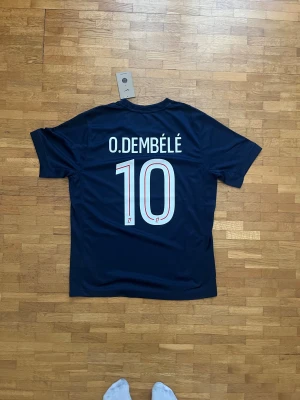 PSG Dembélé #10 Nike fotbollströja M - Säljer en officiell Paris Saint-Germain matchtröja med O. Dembélé och nummer 10 på ryggen. Tröjan är marinblå med röda och vita detaljer, har korta ärmar och är tillverkad i Nike Dri-FIT material. Snyggt geometriskt mönster framtill och PSG-logga på bröstet.