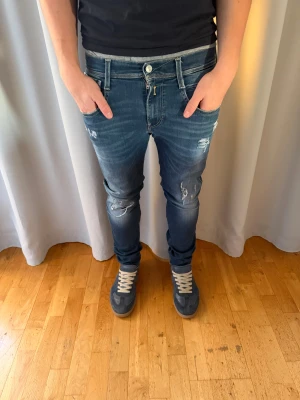 Replay Anbass blå slim jeans med slutningar - Snygga Replay Anbass jeans i blå tvätt med slitna detaljer och slim fit-passform. Klassisk femficksmodell, med dragkedja och knapp framtill. Jeansen har coola slitningar på låren och knäna, samt ljusare partier för en trendig look.