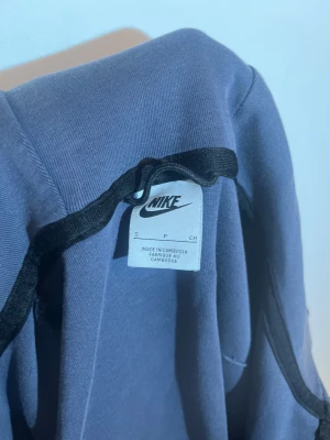 Blå Nike hoodie med dragkedja - Snygg blå hoodie från Nike i storlek S med svart dragkedja framtill och en extra dragkedja på ärmen. Tröjan har huva och ett stilrent Nike-logotryck på bröstet. Materialet är mjuk bomull och polyester, perfekt för chill eller träning.