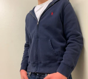 Mörkblå hoodie från Polo Ralph Lauren - En riktigt snygg mörkblå hoodie från Polo Ralph Lauren. Tröjan har huva, dragkedja framtill och två stora fickor. Tröjan är smått tvätt skadad men ser bra ut ändå. 