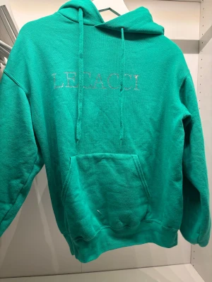 Grön hoodie - Säljer en grön hoodie från LECACCI med strassdetaljer på bröstet och en stor glittrig fjäril på ryggen. Tröjan har huva med snörning, magficka och långa ärmar. Perfekt för dig som gillar statement-plagg och snygga detaljer.
