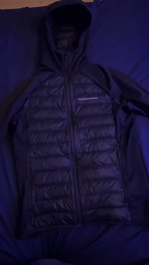 Svart dunjacka Peak Performance - Svart dunjacka från Peak Performance med huva och hel dragkedja. Jackan har quiltad framsida och baksida, samt släta svarta ärmar. Perfekt för kyliga dagar och har en clean, sportig look. Loggan är broderad på bröstet. Kan gå ner i pris 