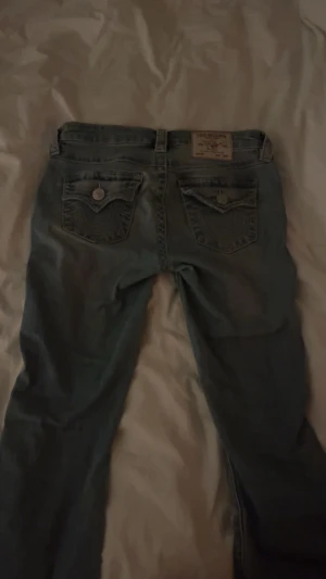 True Religion Becca Bootcut Jeans - Säljer ett par snygga blå jeans från True Religion, modell Becca Mid Rise Bootcut. Jeansen har klassiska fickor med lock och silverfärgade knappar, samt tydliga sömmar och logopatch bak. Perfekt passform med bootcut. Nästan oanvända