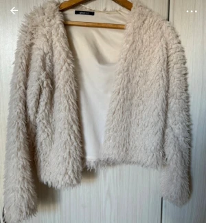 Beige pälsjacka från Ginatricot - Säljer en fluffig och mjuk beige pälsjacka från Ginatricot. Jackan är croppad med långärmade ärmar och har en öppen front utan knappar eller dragkedja. Perfekt för dig som vill ha en cozy och trendig look.