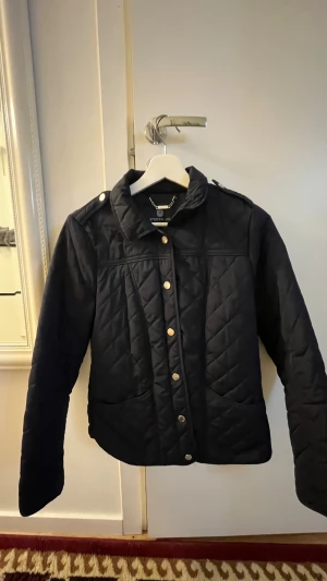 Svart quiltad jacka från Stockh LM - Snygg svart quiltad jacka från Stockh LM i storlek 36. Jackan har guldfärgade knappar, axelklaffar och två stora fickor framtill. Klassisk krage och långärmad modell, perfekt för kyligare dagar. Stilsäker och enkel att matcha till olika outfits.