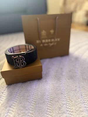Svart Burberry bälte med metallspänne - Snyggt svart bälte från Burberry med klassiskt rutigt innerfoder och ett stort metallspänne med Burberrys ikoniska B-logga. Bältet har en stilren design och passar perfekt till jeans eller kostymbyxor. Kommer i originalförpackning.