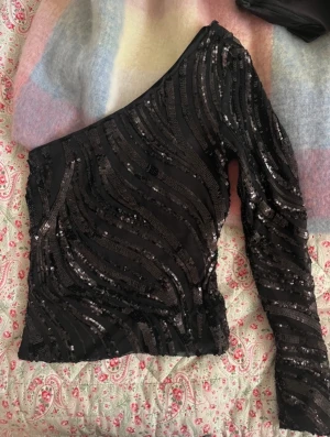 Svart paljett topp från NLY One - Säljer en svart oneshoulder topp från NLY One i storlek 34. Toppen har ett glittrigt paljettmönster i vågiga linjer och en lång ärm. Perfekt till fest