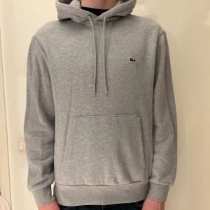 Grå hoodie från Lacoste med huva - Snygg grå hoodie från Lacoste med klassisk krokodillogga på bröstet. Tröjan har huva med snörning, stor magficka och ribbade muddar vid ärmslut och nederkant. Perfekt för chill dagar och enkel att matcha med jeans eller joggers. Krymt lite i tvätten så den har något kortare ärmar