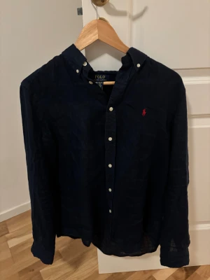 Mörkblå linneskjorta Polo Ralph Lauren XL - Snygg mörkblå skjorta från Polo Ralph Lauren i storlek XL, tillverkad i linne. Klassisk button-down krage, vita knappar och ikonisk röd broderad logga på bröstet. Långärmad och loose fit, perfekt för en chill och stilren look.