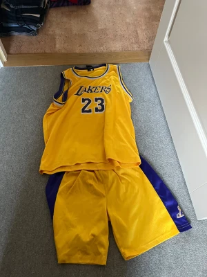 Gul Lakers James 23 baskettröja - Säljer en gul baskettröja och shorts från Lakers med nummer 23 och namnet James på ryggen. Tröjan är ärmlös med blå och vita detaljer vid hals och ärmhål. Materialet är 100% polyester, perfekt för sport och träning. Klassisk Lakers-stil med tryck framtill och bak.