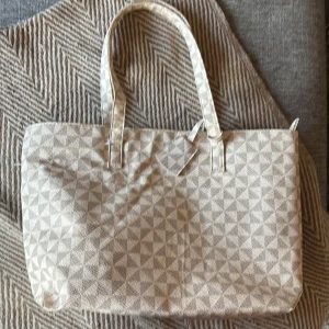 Beige shoppingväska med geometriskt mönster - Stor och rymlig shoppingväska i beige tyg med geometriskt mönster. Väskan har två långa handtag och ett stort huvudfack med innerficka som stängs med dragkedja. Perfekt för att bära med sig allt man behöver under dagen.