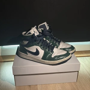 Nike Air Jordan 1 Mid blå/grön - Nike Air Jordan 1 Mid sneakers i blått, grönt och vitt med klassisk Jordan-logga på sidan. Skorna har snörning, rund tå och platt sula. Tillverkade i skinn med snygga paneler och perforerad tå. Levereras med originalkartong och extra vita skosnören.