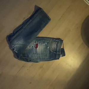 Replay Anbass blå slitna jeans - Säljer ett par Replay Anbass jeans i blå tvätt med slitningar och trasiga detaljer, inklusive en röd patch på låret. Jeansen har klassisk femficksdesign, knappgylf och raka ben. Perfekt för dig som gillar en edgy och avslappnad stil.