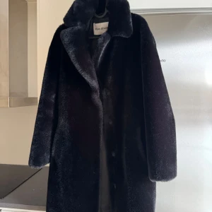 Svart pälsjacka från Stand Studio - Säljer en svart pälsjacka från Stand Studio med klassisk krage och knappar framtill. Jackan har en rak och oversized passform med långa ärmar och mjukt, fluffigt material som ger en lyxig vibe. Perfekt för dig som vill ha en statement piece i garderoben.
